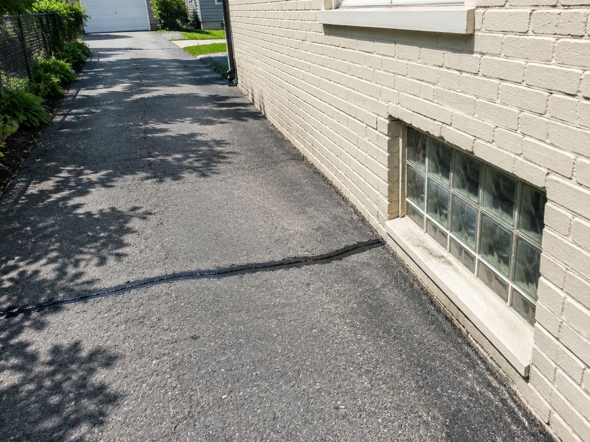Beside a glass-block basement window in a cream brick wall, fresh filler seals a crack angling across a narrow, shaded side drive