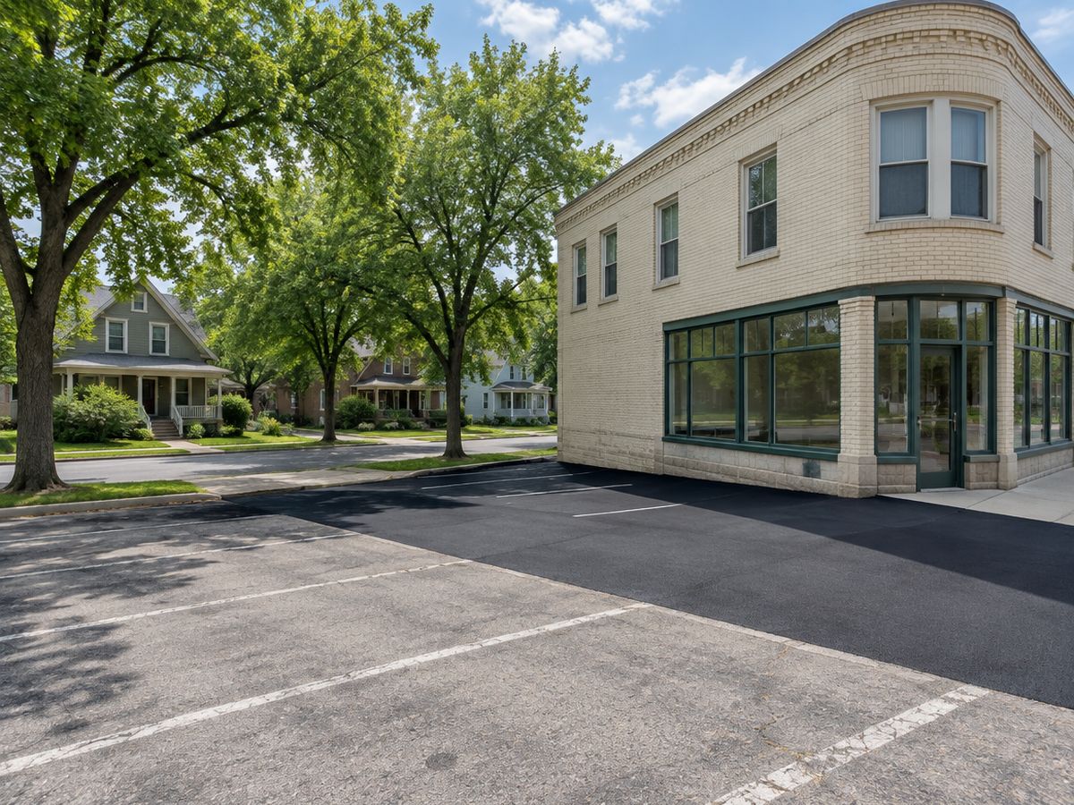 Green-framed windows wrap a cream brick corner storefront, its lot fresh black with new lines beside the building and still faded in the foreground